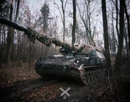 На фото серед зимового лісу стоїть гусенична самохідна артилерійська установка темного кольору, замаскована сітками й гіллям по всьому корпусу та вздовж довгого ствола гармати. Машина розташована на вкритій листям ґрунтовій дорозі між деревами, навколо — похмуре небо та голі стовбури, що підкреслюють приховану бойову позицію техніки