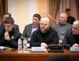На фотографії зображено групу чоловіків, які сидять за столом під час офіційної наради, при цьому на ідентифікаційних картках видно імена «Микола Якович Нарижний» (центральний чоловік з бородою) та «Василь Іванович Книга» (чоловік праворуч у чорному піджаку)
