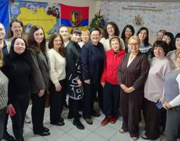 Групове фото учасників заходу соціальної сфери Луганщини в укритті: декілька десятків людей стоять щільним колом у приміщенні, на задньому плані – стінди з логотипами, прапорами та мапою України, поруч новорічна ялинка, усі одягнені в повсякденний одяг і дивляться в бік камери