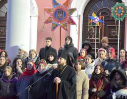 Група людей різного віку, серед яких багато молоді в національному українському одязі та зимових куртках, зібралася на вулиці перед будівлею з білими колонами та арковими дверима. У центрі композиції — чоловік у чорному церковному вбранні та головному уборі, схожому на священника. Деякі учасники тримають великі, багато прикрашені різдвяні зірки-вертепи, що є традиційним атрибутом українського різдвяного дійства — вертепу