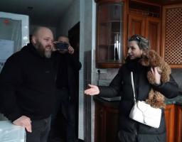 На фотографії зображено людей у приміщенні, схожому на кухню, де жінка тримає на руках коричневого пуделя, а поруч стоїть чоловік у чорній кофті, тоді як інший чоловік на задньому плані знімає сцену на камеру