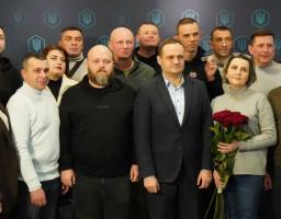 Групове фото колективу робочої команди, ймовірно, з Координаційного центру допомоги ВПО у Кропивницькому. На темному фоні з повторюваними логотипами тризуба та написом "НА" (можливо, "НАШ" чи "НАЦІЯ") позують понад 20 осіб різного віку, переважно чоловіків середнього віку з короткими зачісками та бородами, а також кілька жінок. Центральна постать — чоловік у темному костюмі з синьою сорочкою тримає букет червоних троянд, інші одягнені в повсякденний одяг: светри, куртки, худі