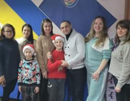 У приміщенні на фоні великого синьо-жовтого прапора та ще одного синього прапора з емблемою стоїть група дорослих і двоє дітей у новорічних шапках. Усі позують щільним колом, деякі тримають дітей за плечі чи за руки, створюючи атмосферу дружньої святкової зустрічі.