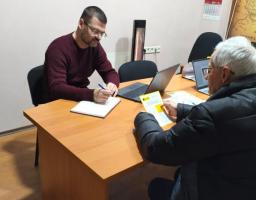 Наданий альтернативний опис до фото: два чоловіки сидять за офісним столом, один з яких пише в блокноті, а інший розглядає документи
