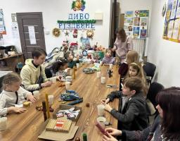кімната з двова великим вікнами, посред кімнати стоїть великий дерев'яний стіл, за яким зібралося багато дітвори. На столі стоять смаколики, різдвяні свічки.