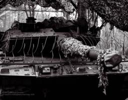На чорно-білій фотографії зображено передню частину танка, ймовірно Stridsvagn 122 або Leopard 2, який належить 121-й окремій механізованій бригаді Збройних Сил України, оснащений додатковим динамічним захистом та камуфляжем, і розташований у лісовій місцевості