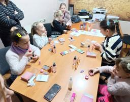 Група дітей та дорослих бере участь у майстер-класі з косметики та догляду за собою, сидячи за офісним столом, заповненим різноманітними продуктами та інструкціями