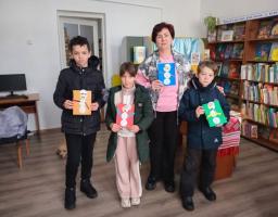 Чотири дитини та доросла жінка, ймовірно, вчителька чи бібліотекарка, стоять разом у бібліотеці та гордо демонструють свої зимові вироби-аплікації із зображеннями сніговиків. Цей альтернативний текст описує основну сцену, включаючи кількість людей, місце дії та предмети, які вони тримають.