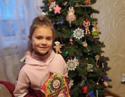 На фотографії зображено молоду дівчину, яка посміхається, тримаючи святкову коробку з зображенням Діда Мороза, стоячи поруч із прикрашеною ялинкою та лялькою сніговика, що створює радісну новорічну або різдвяну атмосферу