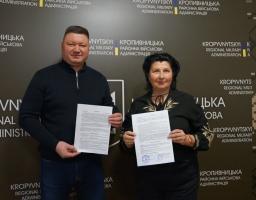 Двоє людей, чоловік та жінка, позують для офіційного фото в приміщенні з брендуванням Кропивницької районної військової адміністрації, тримаючи в руках підписані документи