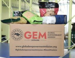 Коричнева картонна коробка з логотипом Global Empowerment Mission Ukraine та контактною інформацією наповнена різними продуктами харчування, такими як пакети з макаронами та крупами, що символізує гуманітарну допомогу від організації.