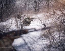 На фотографії зображено українського військовослужбовця, який перебуває на бойовій позиції в сніговому лісі або полі, цілиться з автомата та ховається серед гілок, що свідчить про виконання бойового завдання в зимових умовах.