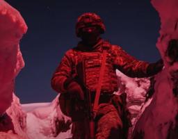 Український військовий у повному спорядженні та шоломі стоїть у засніженому окопі вночі, освітленому насиченим червоним світлом, яке підкреслює мілітаристську атмосферу.