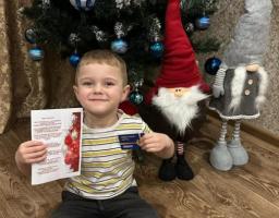 Щасливий маленький хлопчик сидить на дерев'яній підлозі перед різдвяною ялинкою, прикрашеною синіми кульками, та двома декоративними гномами, гордо демонструючи білу листівку та синю картку, які він тримає в руках