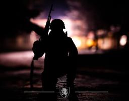 на темному тлі, освітленому лише розмитими вогнями міста або військової бази, зображено силует озброєного солдата, який стоїть на коліні з автоматом, а внизу розташований герб або емблема військової частини, що підкреслює урочистість моменту