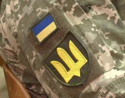 Крупним планом зображено рукав військової форми зі сучасним піксельним камуфляжем, на якому розташовані дві нашивки: менша прямокутна із синьо-жовтим прапором України та більша емблема тризуба золотистого кольору на темному щиті