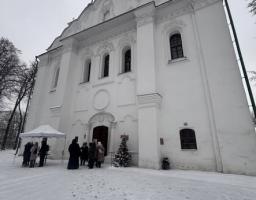 Біла церква з зеленим куполом стоїть на засніженій землі, де група людей зібралася біля входу поруч із маленькою прикрашеною ялинкою та білим наметом.