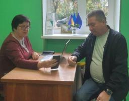 жінка в окулярах та чоловік у куртці, сидять за дерев'яним столом у приміщенні з яскраво-зеленими стінами та обговорюють щось, дивлячись на смартфон жінки, тоді як на підвіконні видніються маленькі українські прапори, що свідчить про офіційну або соціальну зустріч в Україні.