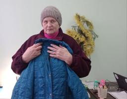 жінка середнього віку в бордовій куртці та сірій в'язаній шапці стоїть у приміщенні зі світло-зеленими стінами, тримаючи перед собою синій стьобаний плащ, а на задньому плані видніється декоративна ялинка та робочий стіл