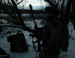 Український військовослужбовець у повному бойовому спорядженні, включаючи шолом, тактичний жилет і рюкзак, сидить на колінах на засніженій землі в густих заростях, тримаючи в руках автоматичну зброю з прикріпленим великим глушником, на тлі темного зимового пейзажу