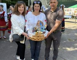 Три людини — дві жінки та один чоловік — позують разом, тримаючи плетений кошик із різноманітними предметами, на тлі відкритої сцени та дерев