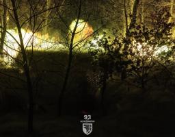 Темна нічна сцена в лісі із силуетами голих дерев і вічнозелених кущів, освітлена яскравим жовтим світлом на задньому плані, із нашивкою 93-ї бригади «Холодний Яр» унизу по центру