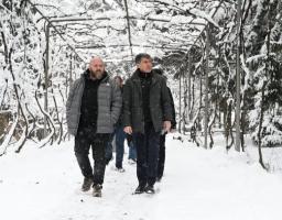 Два чоловіки в зимовому одязі йдуть засніженим проходом, утвореним аркою з покритих снігом гілок, із додатковими людьми та деревами на задньому плані.