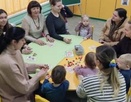 Група дорослих і дітей сидить навколо зеленого столу в класі або дитячому закладі, де розкладені іграшки або навчальні матеріали для спільної гри
