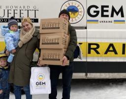 українська сім'я з двома дітьми стоїть на засніженій землі біля вантажівки з логотипами Howard G. Buffett Foundation та GEM Ukraine, отримуючи пакунки гуманітарної допомоги від цих благодійних організацій