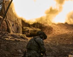 український солдат на передньому плані, який спостерігає, як бронетранспортер стріляє, створюючи яскравий вибух і клуби диму з вогнем у прихованій вогневій позиції, ймовірно, під час російсько-української війни.