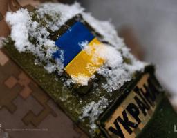 Крупний план військової форми з нашивкою українського прапора та написом «УКРАЇНА», присипаних снігом, з додатковим логотипом та текстом дати в нижньому лівому куті.