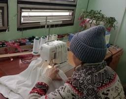 Людина в синій в'язаній шапці та светрі з норвезьким візерунком сидить за дерев'яним столом і шиє на білій швейній машині білу тканину, з додатковими нитками та рослинами на фоні