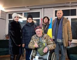 зображено українського волонтера дядька Гришу (Григорія Янченка) в інвалідному візку, який сидить у приміщенні з групою інших людей, зокрема жінками у зимовому одязі та чоловіком у пальті, тримаючий невеликий синьо-жовтий прапор