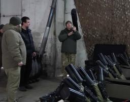 На зображенні група з трьох чоловіків у зимовому та військовому одязі перебуває в приміщенні, схожому на склад або майстерню. На передньому плані видно кілька рядів чорних ракетних або протитанкових комплексів, акуратно складених у стелажах.