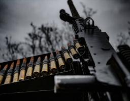 Великокаліберний кулемет розташований на передньому плані, поруч із стрічкою патронів, готових до використання, на тлі сірого неба та розмитих дерев