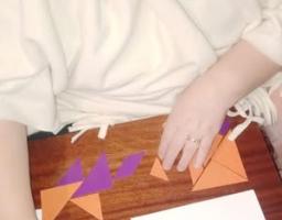 Зображення показує людину в окулярах, яка сидить за дерев'яним столом і складає фігури з оранжевих та фіолетових частин головоломки танграм, поруч лежить аркуш із прикладами та написом TANGRAM