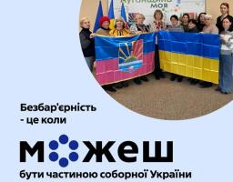 Група з приблизно десяти людей, переважно жінок, стоїть у приміщенні та тримає розгорнутими два великі прапори: праворуч — синьо-жовтий державний прапор України, ліворуч — інший прапор із синьо-рожевими горизонтальними смугами та гербом. На задньому плані видно банер з написом «Луганщина моя». Великий текст на передньому плані зображення говорить: «Безбар'єрність - це коли можеш бути частиною соборної України»