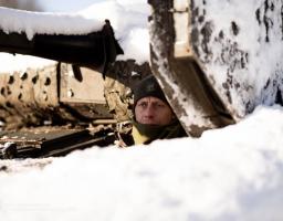 Крупний план українського солдата в зеленій шапці та шарфі, який визирає із засніженої військової техніки на зимовому полі