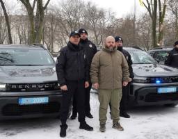 Група чоловіків у поліцейській формі та цивільному одязі, включно з Оксеном Лісовим (другий праворуч), стоять біля нових службових автомобілів Renault Duster. Ці автомобілі (з номерними знаками, що містять цифри 2088 та 2089) були передані підрозділам Національної поліції України, зокрема для поліцейських офіцерів громад. Подібні передачі автомобілів регулярно відбуваються в різних регіонах України, включаючи прифронтові області, для покращення матеріально-технічного забезпечення правоохоронців.