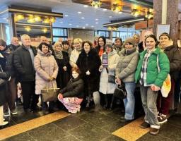 Група з близько двох десятків людей різного віку в зимовому одязі зібралася в приміщенні з темною плиткою на підлозі та декоративними світильниками на стелі