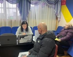 На передньому плані сидить лисий чоловік у темній куртці, який тримає документ і чекає. За столом праворуч від нього сидить жінка за ноутбуком, перед якою стоїть табличка з написом «ЄДИНЕ ВІКНО». Праворуч на синьому стільці сидить літня жінка у фіолетовій куртці. На задньому плані видно вікно із синіми шторами та на стіні — частину українського прапора та прапора іншого регіону