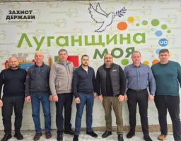 На зображенні група з шести чоловіків стоїть у приміщенні перед великим рекламним банером. На банері написано «Луганщина МОЯ» та «ЗАХИСТ ДЕРЖАВИ», а також зображено голуба миру та різнокольорові кола. Чоловіки одягнені у повсякденний одяг, включаючи светри, джинси та жилети