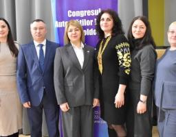 групове фото шістьох людей (три жінки та троє чоловіків), які позують стоячи в приміщенні перед фіолетовим банером і сірою шторою. Люди одягнені в офіційний або напівформальний одяг. Серед них двоє чоловіків у костюмах, жінка в центрі в сірому костюмі, жінка в чорній сукні з традиційною вишивкою, жінка в бежевій сукні та ще одна жінка в синьому одязі. На задньому плані видно частину банера з текстом румунською мовою, зокрема слова "Congresul" та "Autotilor"