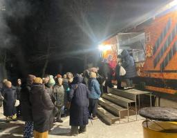 На зображенні показана група людей на вулиці вночі, які стоять біля великого оранжевого автомобіля, можливо, фудтрака, на снігу. Люди одягнені в зимовий одяг.