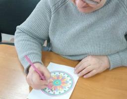 Людина в окулярах і сірому светрі сидить за дерев'яним столом і розфарбовує малюнок мандали рожевим олівцем. На столі видно розсипані кольорові олівці та відкриту упаковку олівців українського бренду «Барвінок» на 12 кольорів