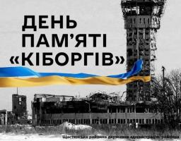 Чорно-біле фото зруйнованого будинку та диспетчерської вежі на тлі зимового пейзажу, з великим текстом «ДЕНЬ ПАМ'ЯТІ «КІБОРГІВ»» угорі та синьо-жовтим українським прапором, що майорить через центр зображення, а також дрібним написом «Щастинська районна державна адміністрація районна військова адміністрація» внизу