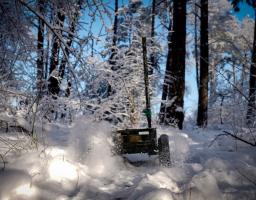 На зображенні показано невеликий військовий наземний роботизований комплекс (НРК) на колесах зеленого кольору, який активно рухається глибоким снігом у засніженому лісі, створюючи бризки снігу. Такі роботи використовуються українськими військовими для різних завдань, включаючи розвідку та евакуацію