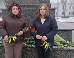Дві жінки стоять поруч із гранітним пам'ятником або меморіалом у засніженому парку взимку. Вони тримають букети квітів (деякі червоні гвоздики, деякі жовто-сині квіти), перев'язані синьо-жовтими стрічками, що символізують український прапор. На землі біля основи пам'ятника також лежать інші букети квітів та стрічки.