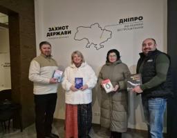 На зображенні четверо людей, троє з яких тримають книги, позують у приміщенні біля стіни з картою України та написами українською мовою: «ЗАХИСТ ДЕРЖАВИ», «ДНІПРО», «ВІД БЕЗПЕКИ ДО ПРОЦВІТАННЯ» та «ЗОЛОТО НАШ». Люди посміхаються і одягнені в повсякденний одяг, один із чоловіків носить жилет