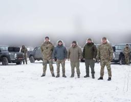 На зображенні група з п'яти чоловіків у військовій формі та зимовому одязі стоїть на засніженому полі перед кількома пікапами сірого кольору. Також на задньому плані видно ще кількох людей і транспортні засоби